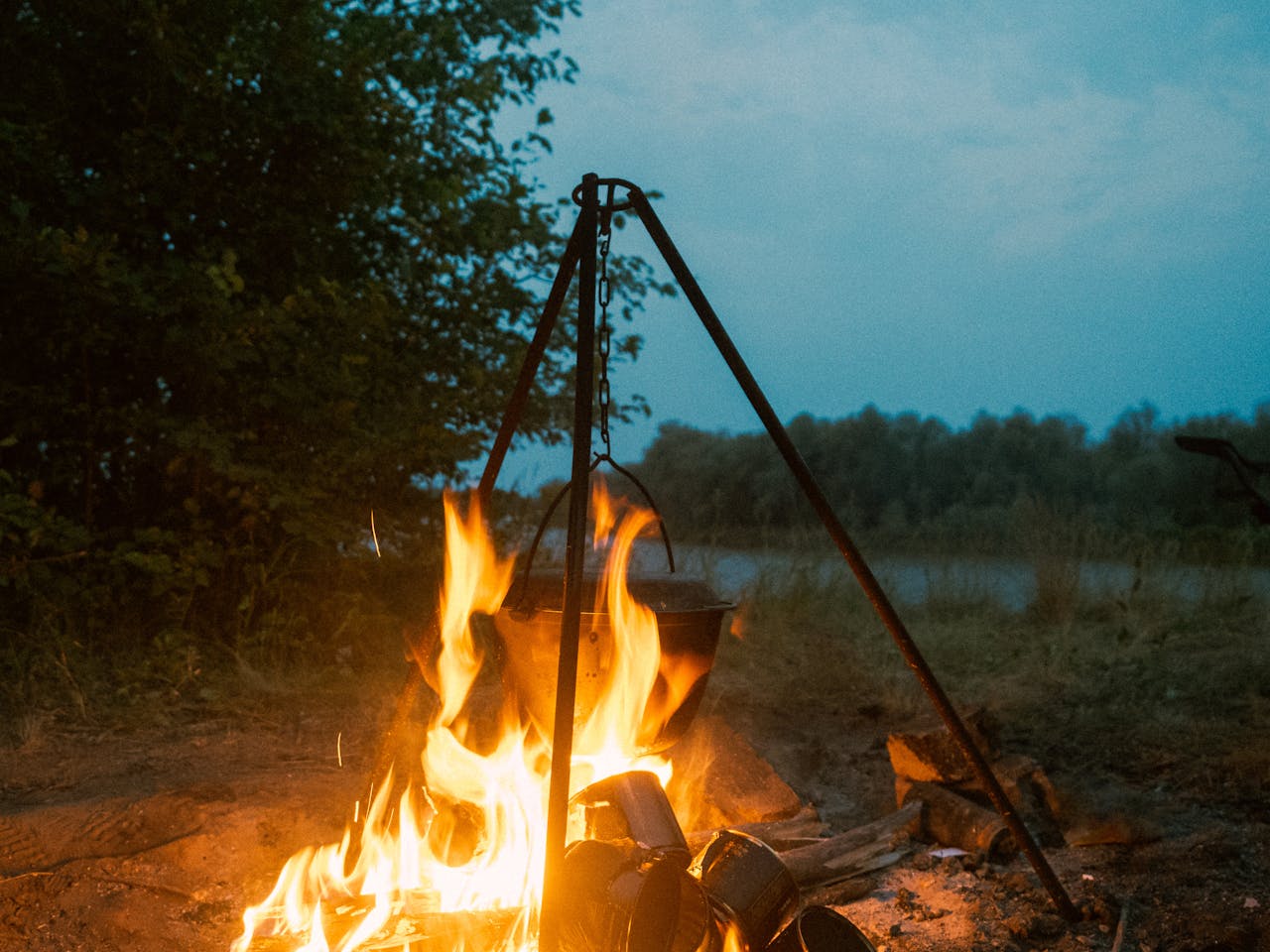 A serene camping scene featuring a burning campfire with a cooking pot outdoors near a lake during evening.